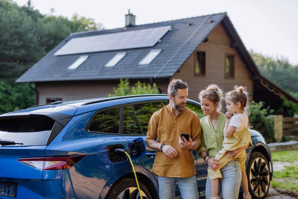 Familie vor ihrem Elektroauto, das vor ihrem Haus mit PV-Modulen auf dem Dach geladen wird.