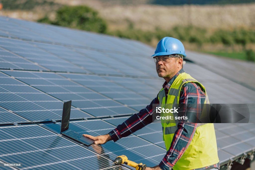An electric engineering mature professional male with a blue helmet and worker west is captured amid work at a photovoltaic farm, laptop in hand and drill. This image signifies the integration of technology and expertise in the maintenance, monitoring, or optimization of the solar panel energy production system. The engineer's use of a laptop suggests engagement in tasks related to data analysis, system monitoring, or perhaps planning for the efficient operation of the photovoltaic farm. This portrayal reflects the intersection of engineering and renewable energy technology in the contemporary pursuit of sustainable power solutions